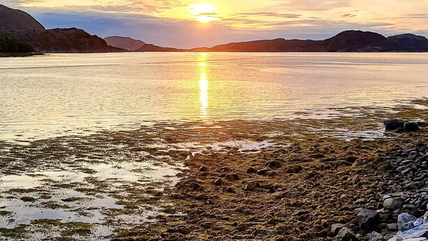 Een rustig meer bij zonsondergang met een rotsachtige oever en heuvels in de verte.