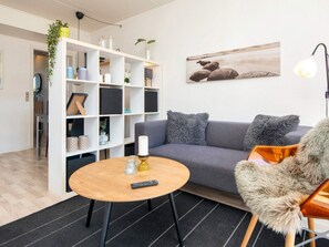 A modern living room with a grey sofa, a round wooden coffee table, a fluffy chair, a white shelving unit, and a painting on the wall. - 2 Person Holiday Home in Ringkobing (Ringkobing)