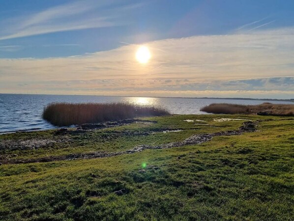 A coastal landscape with a body of water, grassy areas, and a clear sky with the sun low on the horizon. - 2 Person Holiday Home in Ringkobing (Ringkobing)