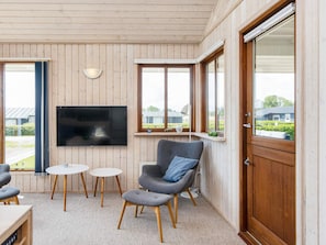 Une pièce avec un téléviseur fixé au mur, un fauteuil gris avec un coussin bleu, deux petites tables rondes et une porte en bois.