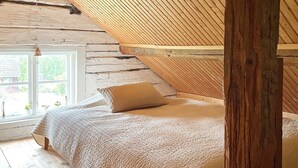 Une chambre avec un plafond en pente, des poutres en bois, une grande fenêtre et un lit recouvert d’une couverture et d’un oreiller beiges.