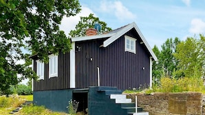 Une petite maison en bois sombre, avec un toit en pente, une cheminée et un petit porche doté de marches menant à l’entrée.