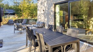 A modern outdoor deck with a wooden table and chairs, surrounded by a brick wall and large windows. - 5 Star Holiday Home in Vestervig (Vestervig)