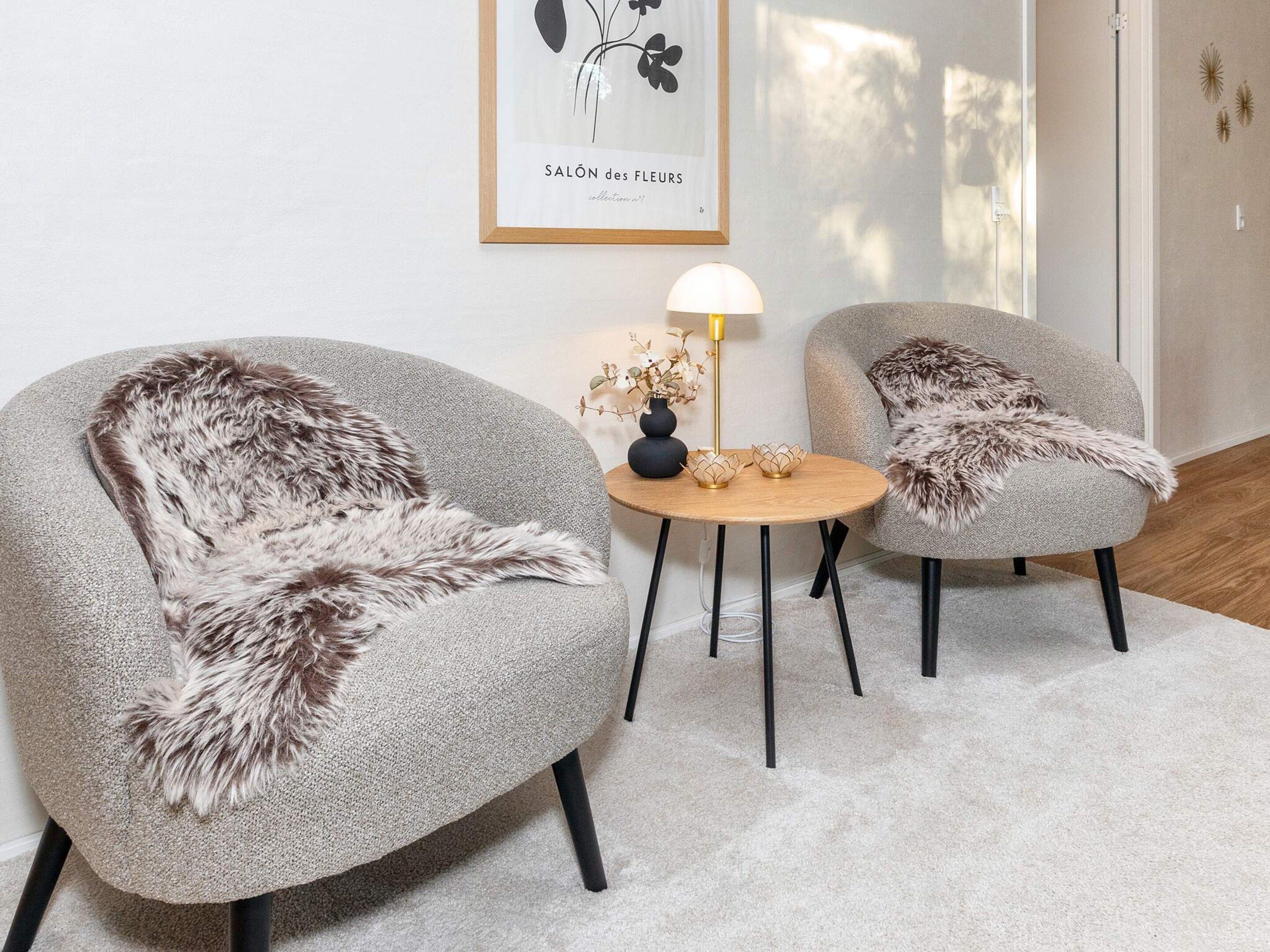 Una habitación con dos sillones grises con almohadones de piel, una pequeña mesa redonda con una lámpara y una obra de arte enmarcada en la pared.