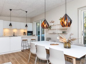 Une cuisine moderne avec des meubles blancs, un îlot central, des luminaires suspendus et un coin repas comprenant une table et des chaises blanches.