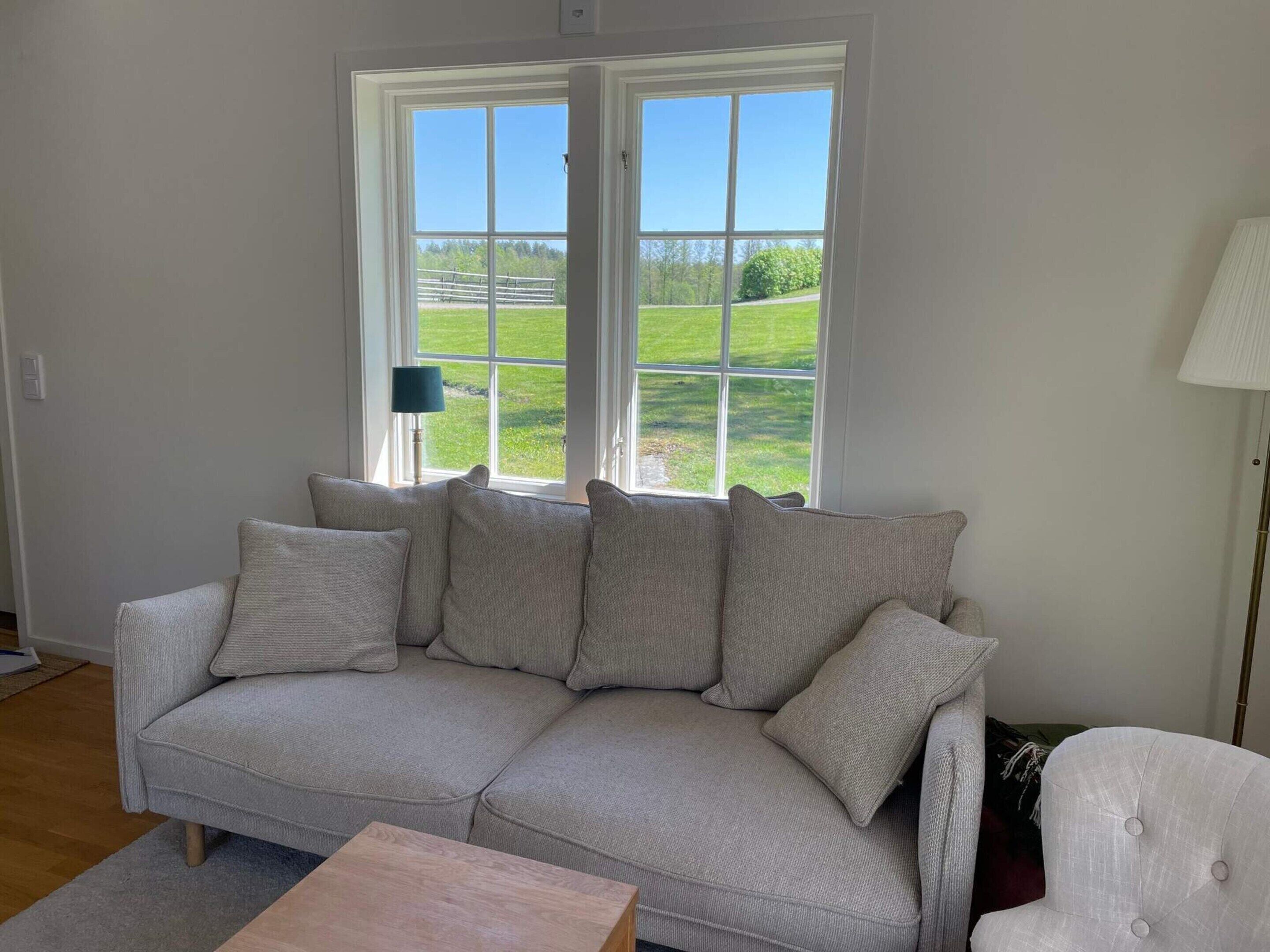 A living room with a beige sofa, a wooden coffee table, and a view of a green landscape through large windows.