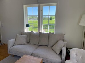 Un salon avec un canapé beige, une table basse en bois et une vue sur un paysage verdoyant, visible à travers de grandes fenêtres.