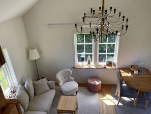 A cozy living room with a sofa, a chair, a small table, a wooden side table, a lamp, a window with a view of greenery, and a chandelier.