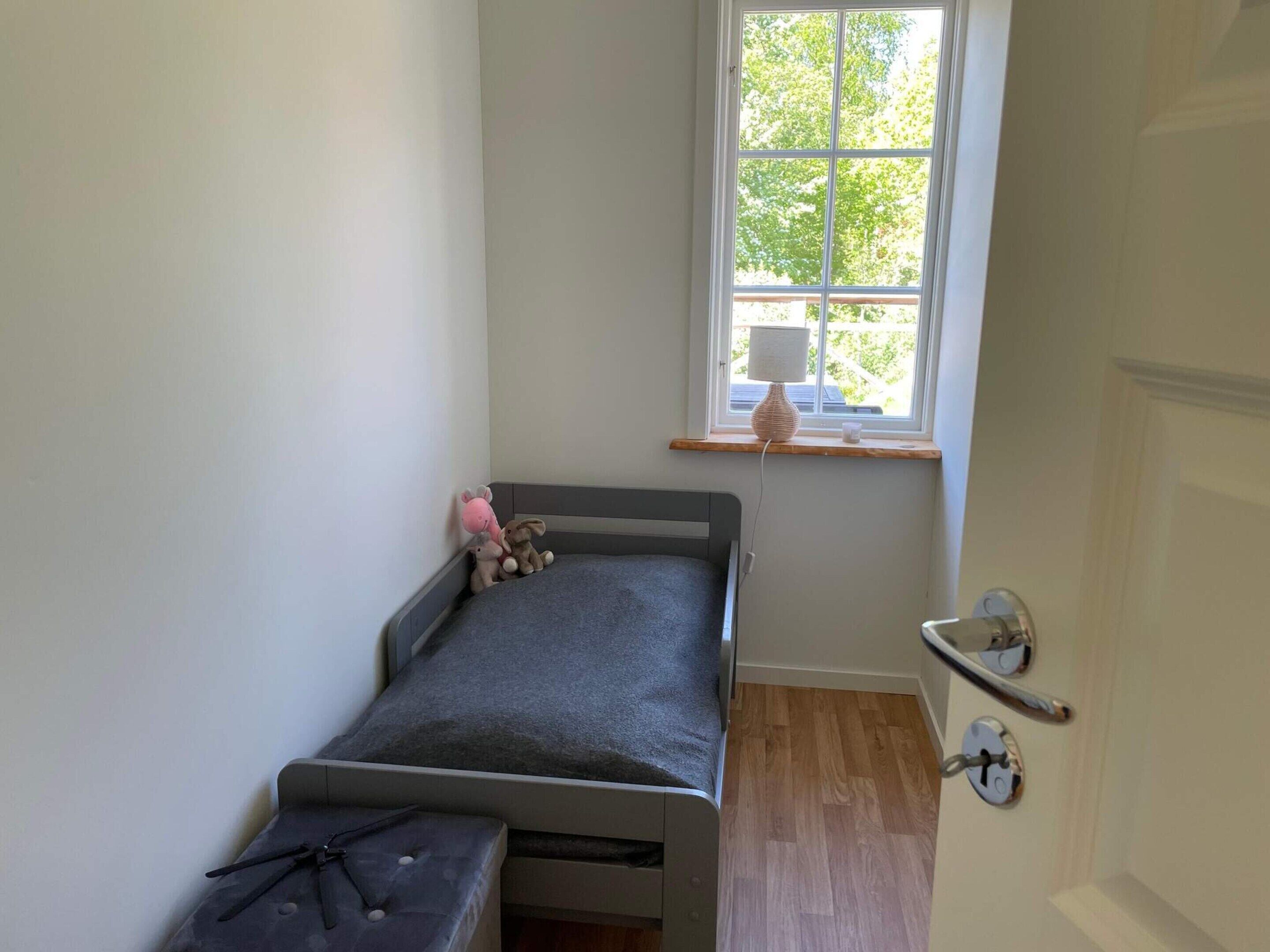 A small, narrow room with a single bed, a window with a view of trees, and a door with a handle.