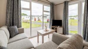 A modern living room with a sofa, a coffee table, and a TV. Large windows offer a view of a caravan park. - Chalet in De Haan Near Sandy Beaches (De Haan)