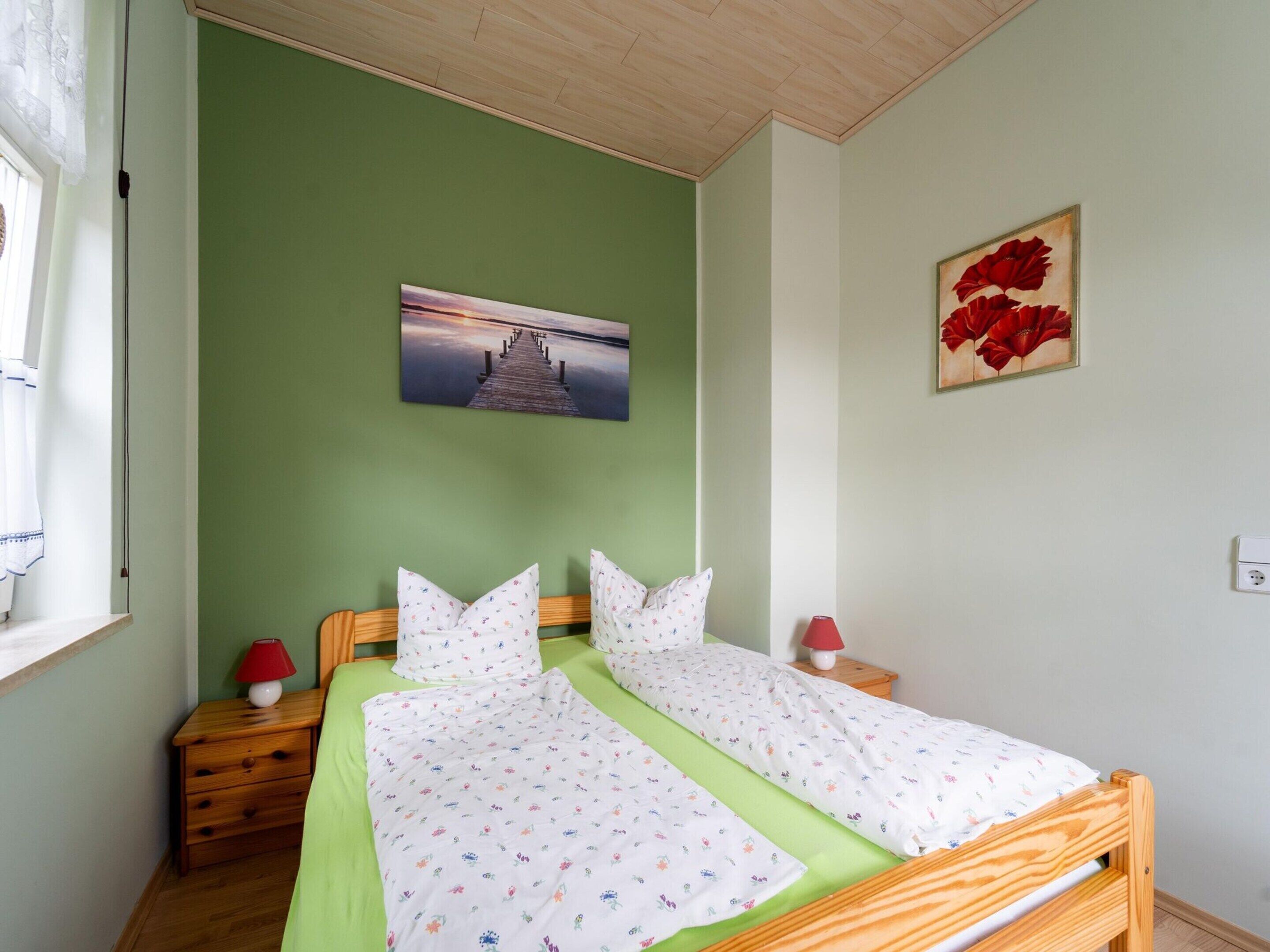 A double bed with floral bedding, a nightstand with a lamp, a painting of a pier, and a red flower painting on the wall.