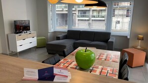 Una sala de estar moderna con un sofá, una mesa de comedor con una manzana verde, un televisor y una ventana con persianas.