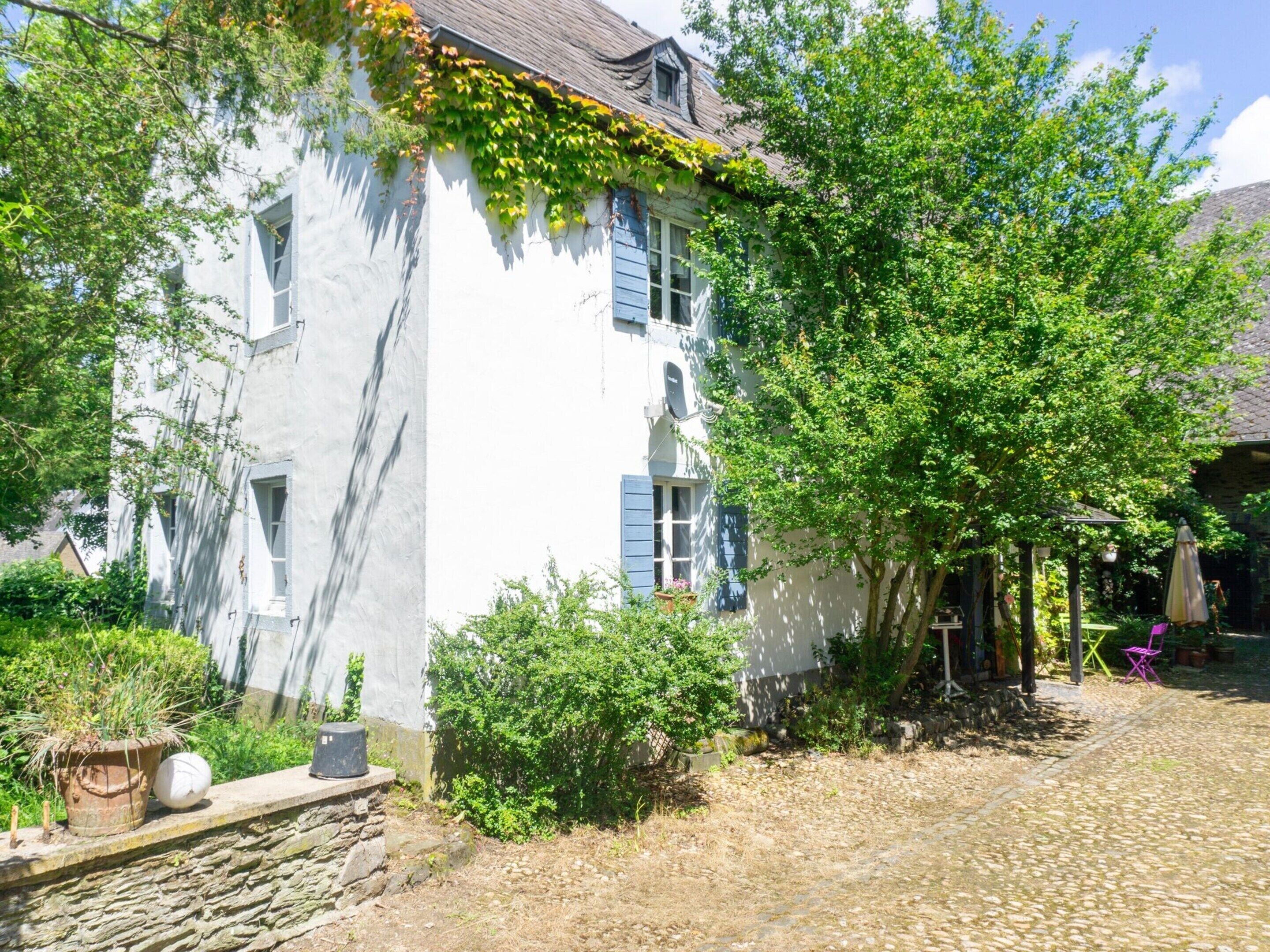 A white building with blue shutters, surrounded by greenery and a cobblestone path.