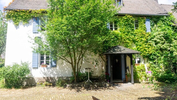 Un bâtiment blanc avec un toit en tuiles, un banc en bois et un petit jardin agrémenté d’un arbre et de plantes en pot.