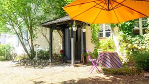 Un espace extérieur abrité avec des sièges, doté d’un grand parasol orange, d’un banc en pierre et d’un petit jardin avec des plantes et un pot de fleurs.