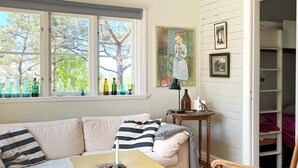 A cozy living room with a white sofa, striped pillows, a wooden coffee table, and a chair. There are two windows with bottles on the sill, a painting on the wall, and a bookshelf in the background. - Attractive Apartment With Terrace-by Traum (Mellbystrand)