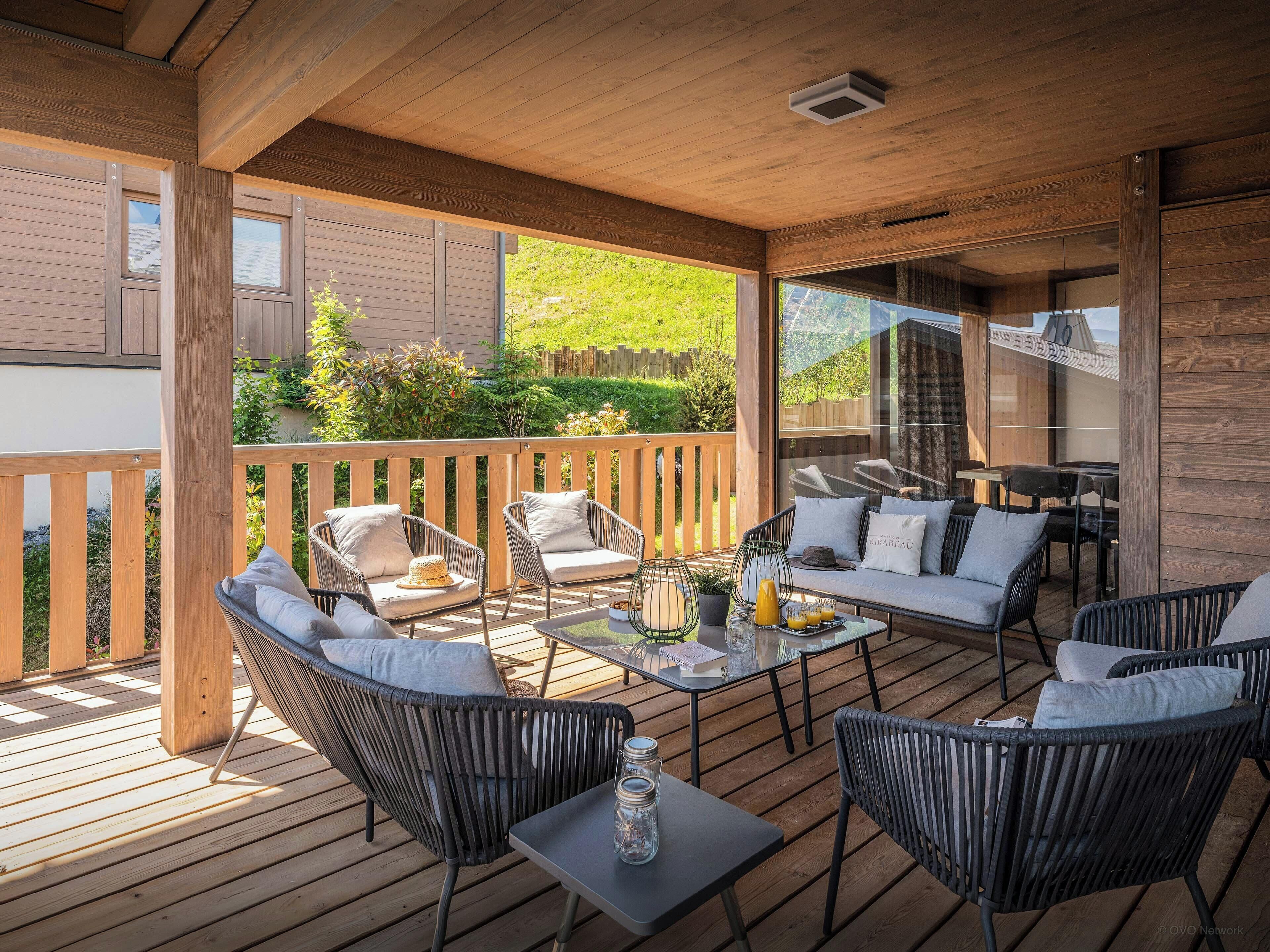 A covered outdoor seating area with wicker chairs, a glass table, and a view of a garden.