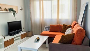 A modern living room with a flat-screen TV, a white coffee table, an orange sofa with multiple cushions, and a grey armchair. - Beachhouse Gran Tarajal 3BR (Gran Tarajal)