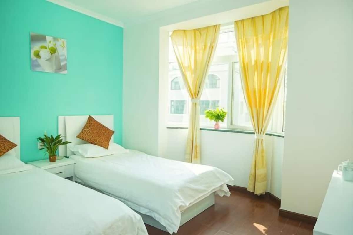Une chambre avec deux lits, une fenêtre ornée de rideaux jaunes, une plante posée sur le rebord de la fenêtre et un tableau accroché au mur.
