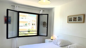 A bedroom with a bed, a nightstand, a lamp, a window with curtains, and a wall with a framed picture. - QUATRINA L - Pretty T2 ideally located (PROPRIANO)