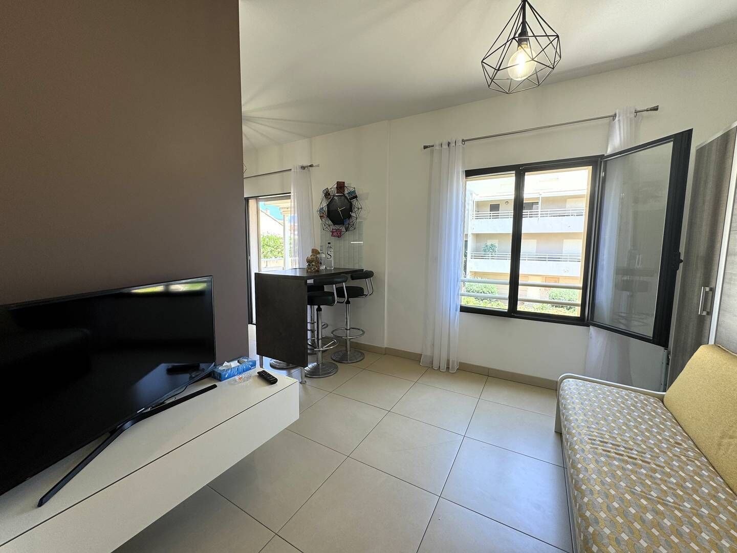 A modern living room with a flat-screen TV, a sofa, a dining area with a table and chairs, a large window with curtains, and a ceiling light fixture.