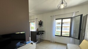 A modern living room with a flat-screen TV, a sofa, a dining area with a table and chairs, a large window with curtains, and a ceiling light fixture. - QUATRINA L - Pretty T2 ideally located (PROPRIANO)