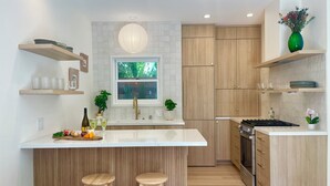 A modern kitchen with wooden cabinets, a central island with stools, a window with a view of greenery, and a hanging light fixture. - Casa Flora, Brand-New Serene Guest House in Los Angeles with WiFi, AC (Los Angeles)
