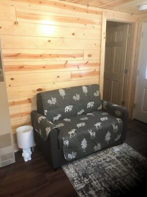 Una habitación con pared de madera, un sofá con funda de motivos naturales, una lámpara blanca y una alfombra estampada.