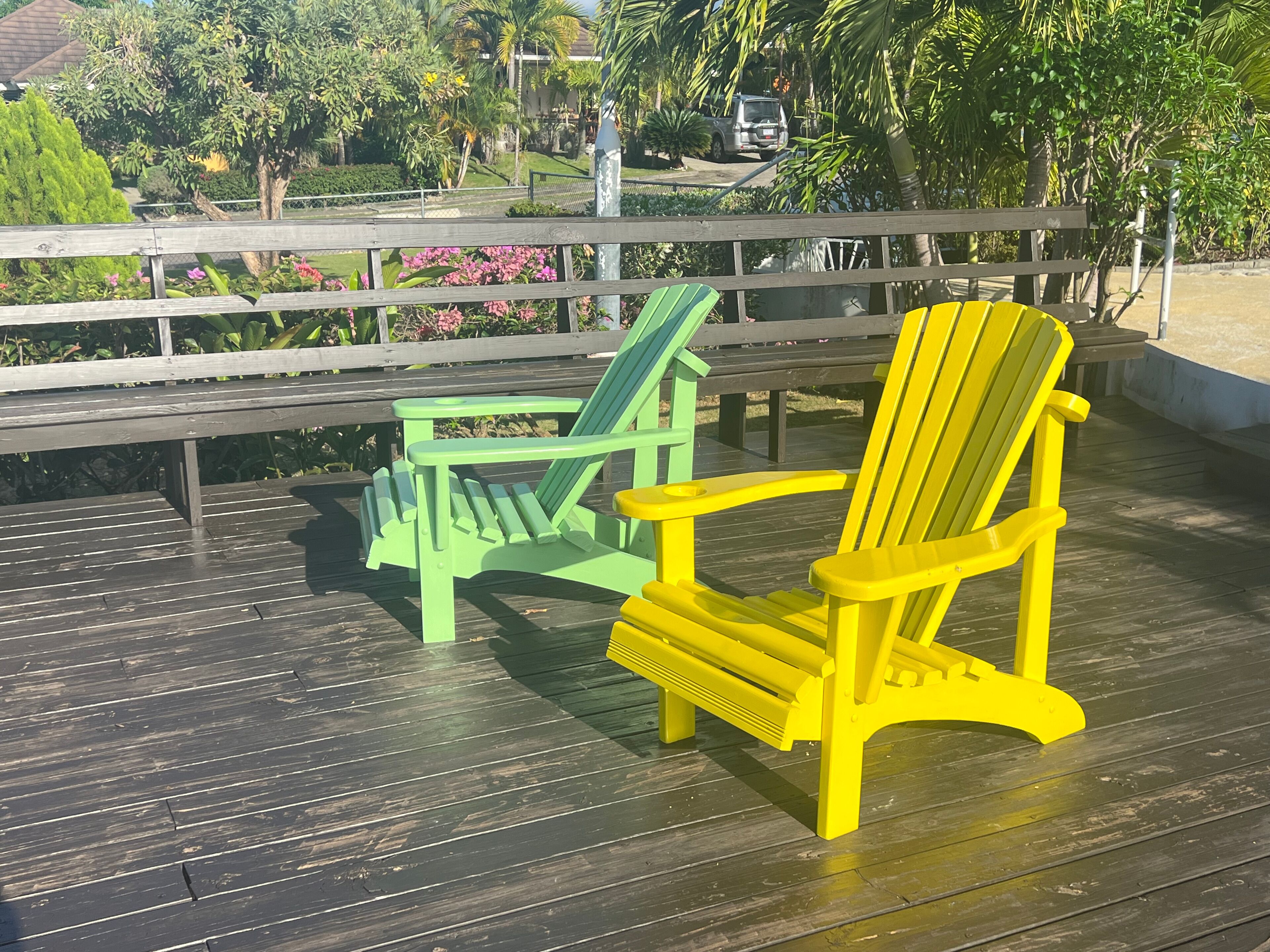 Two Adirondack chairs on a wooden deck with a view of a residential area and greenery.