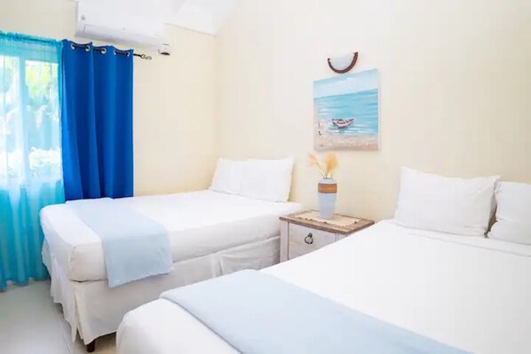 A hotel room with two beds, a small wooden nightstand, a painting of a beach scene, and a window with blue curtains.