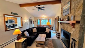 A living room with a fireplace, a ceiling fan, a kitchen area, and a dining area with natural light. - F26 Bretton Woods home steps from the slopes, AC, pool passes, discounted cog tix! (Bretton Woods)
