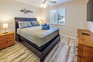 Quarto com cama, mesas de cabeceira, ventoinha no teto, janela com persianas e cômoda de madeira.