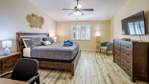 Ein Schlafzimmer mit Bett, Holzkommode, Fernseher, Deckenventilator und Fenster mit Jalousien.