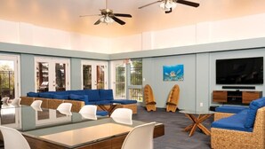 A spacious living area with a glass-top dining table, blue cushioned sofas, wicker chairs, and a flat-screen TV.