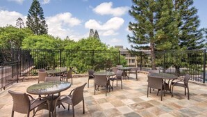 An outdoor patio with round tables and wicker chairs, surrounded by a stone-tiled floor and a metal fence.