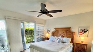 A bedroom with a large bed, a ceiling fan, a nightstand with a lamp, and a view through a sliding glass door.