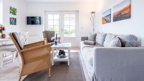A modern living room with a grey sofa, wicker chairs, a white coffee table, and a flat-screen TV mounted on the wall. - Raantem Hüs Whg. 1 - Wattblick. Naturnah. Terrasse (Rantum)