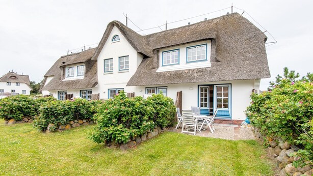 A row of white houses with thatched roofs and blue trim, surrounded by well-maintained hedges and a grassy lawn. - Raantem Hüs Whg. 1 - Wattblick. Naturnah. Terrasse (Rantum)