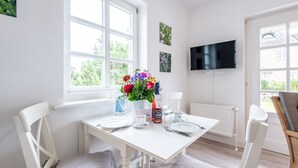 A bright dining area with a white table set for two, a vase of flowers, and a bottle of wine. A flat-screen TV is mounted on the wall above the table. - Raantem Hüs Whg. 1 - Wattblick. Naturnah. Terrasse (Rantum)