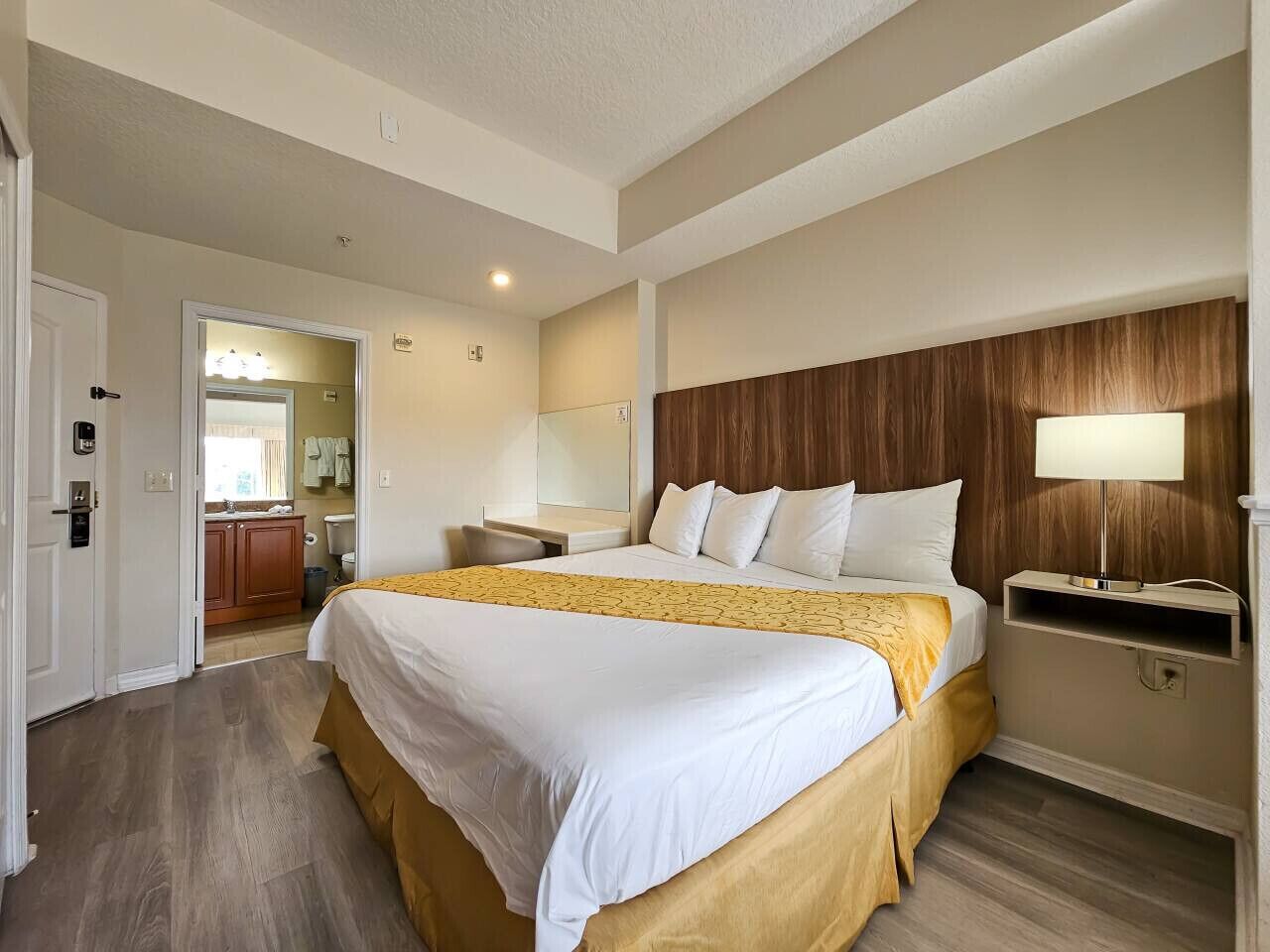 A hotel room with a large bed, a wooden headboard, a bedside table with a lamp, and a bathroom visible through an open door.