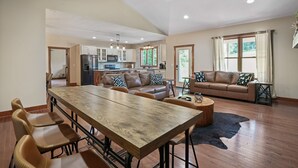 A spacious living area with a dining table, sofas, and a kitchen in the background. - Espolon Ranch | 5BR, Pool, & Pickleball (New Plymouth)
