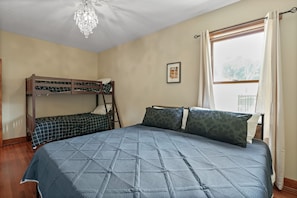 A bedroom with a bunk bed, a large bed with patterned pillows, a chandelier, a window with curtains, and a framed picture on the wall.