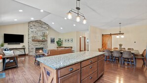 A spacious living area with a stone fireplace, a large TV, and a dining area with a wooden table and chairs. - Espolon Ranch | 5BR, Pool, & Pickleball (New Plymouth)