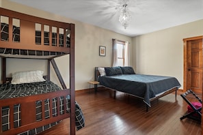 Una habitación con literas, una cama individual, piso de madera, candelabro, ventana con cortinas y una puerta.