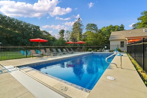 Una piscina rectangular con una escalera de metal, rodeada por una cerca negra y dos sombrillas rojas.
