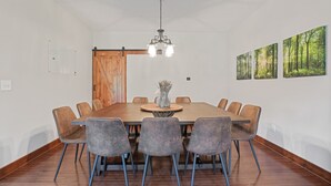 A dining area with a large wooden table, six grey chairs, a hanging light fixture, and three framed forest pictures on the wall. - Espolon Ranch | 5BR, Pool, & Pickleball (New Plymouth)