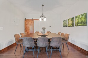 A dining area with a large wooden table, six grey chairs, a hanging light fixture, and three framed forest pictures on the wall. - Espolon Ranch | 5BR, Pool, & Pickleball (New Plymouth)