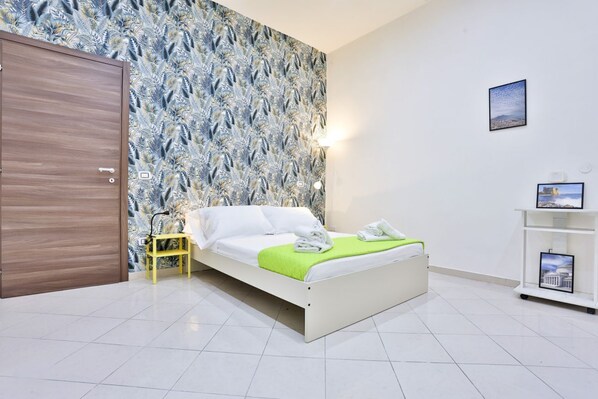 A bedroom with a bed, a nightstand, a wall with a leaf pattern, a door, and a shelf with pictures.