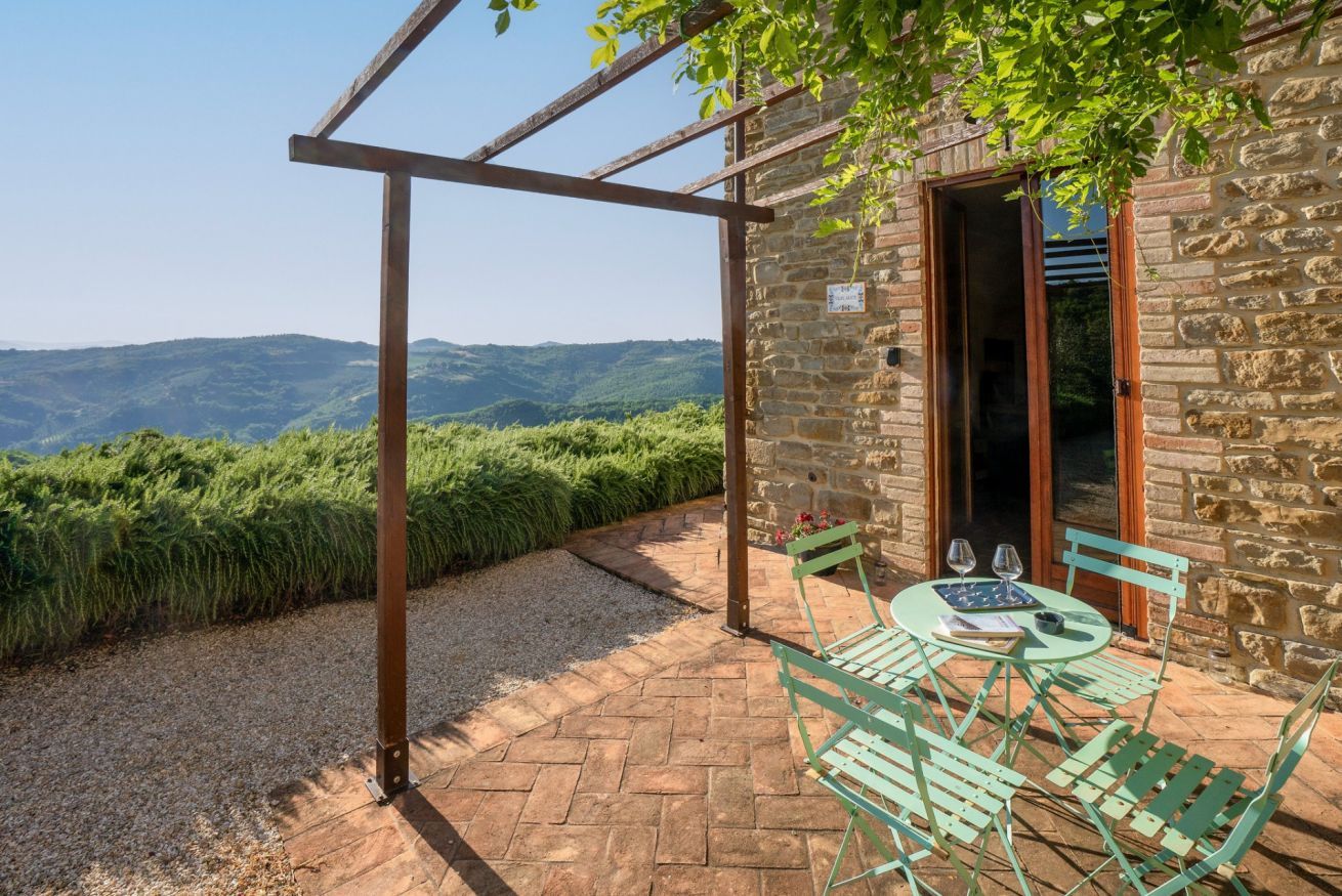 Un edificio de piedra con una pérgola, una mesa con sillas y vistas a las colinas.
