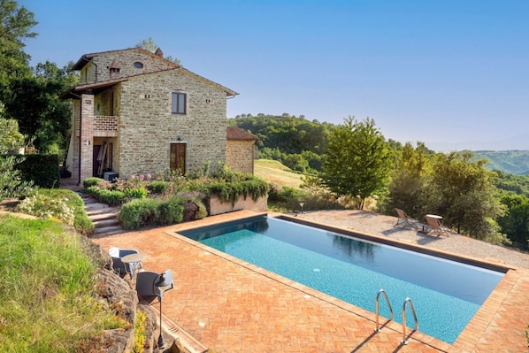 Una casa de piedra con piscina, rodeada de vegetación y colinas.